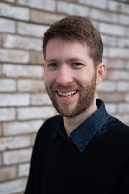 Andrew Hamblin smiling wearing a blue sweater in front of a brick wall